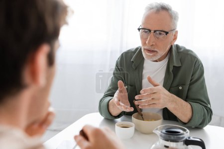 Téléchargez les photos : Senior en lunettes gestuelle lors d'une conversation avec son fils prendre le petit déjeuner dans la cuisine - en image libre de droit