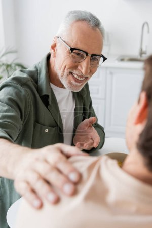 Lächelnder Mann mit Brille, der mit der Hand zeigt und die Schulter seines verschwommenen Sohnes während eines Gesprächs in der Küche berührt