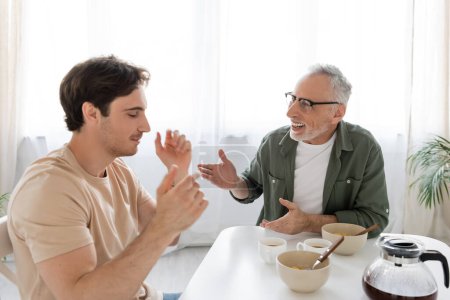 alegre hombre de pelo gris hablando con hijo desanimado durante el desayuno en la cocina