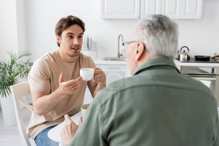man holding coffee cup and pointing with hand during dialogue with dad on blurred foreground
