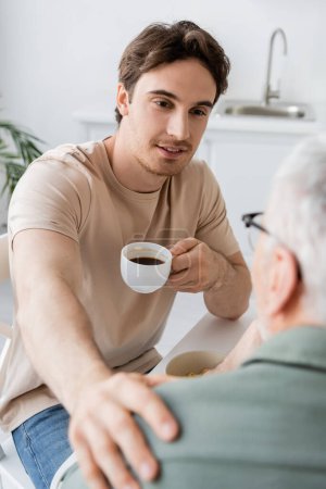 positive young man with coffee cup talking to blurred dad and touching his shoulder in kitchen