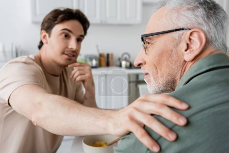 blurred positive man touching shoulder of thoughtful dad looking away during breakfast in kitchen