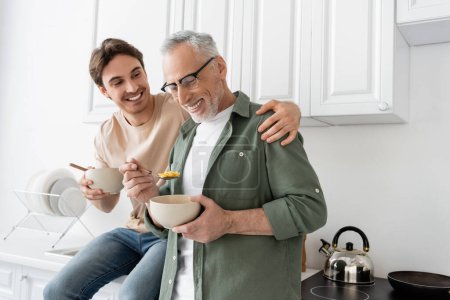 Photo for Happy young man sitting on kitchen worktop and hugging shoulders of smiling dad holding bowl and spoon with corn flakes - Royalty Free Image