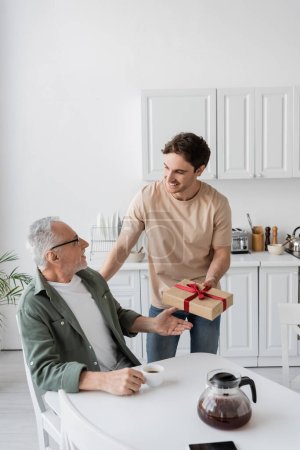 smiling man presenting fathers day gift to pleased dad sitting in kitchen with coffee cup