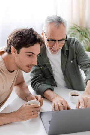 positive mature man pointing at laptop near smiling son holding coffee cup in kitchen