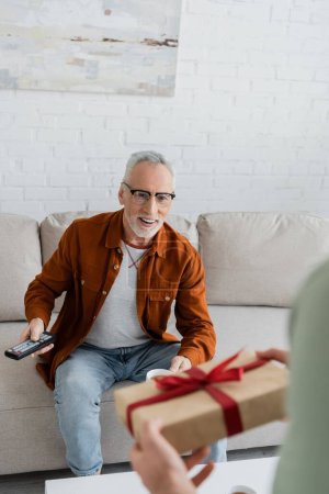 Verschwommener Mann hält Vatertagsgeschenk in der Nähe seines auf Sofa sitzenden Vaters 