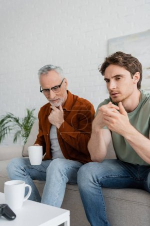 thoughtful mature man with tea cup touching beard while sitting near serious son 