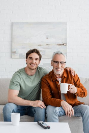 young and happy man embracing shoulders of smiling dad holding tea cup while looking at camera