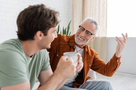 fröhlicher älterer Mann mit Brille gestikuliert und spricht mit erwachsenem Sohn mit Teetasse im verschwommenen Vordergrund