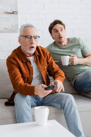 Shocked senior man with remote controller and son with tea cup watching tv at home