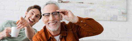 Photo for Cheerful man adjusting eyeglasses near laughing son with tea cup pointing with finger, banner - Royalty Free Image
