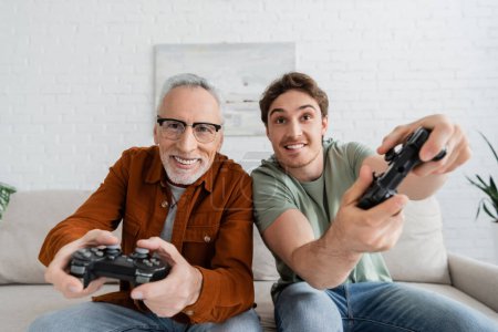 Photo for KYIV, UKRAINE - MAY 11, 2022: excited man and mature cheerful dad in eyeglasses gaming in living room - Royalty Free Image