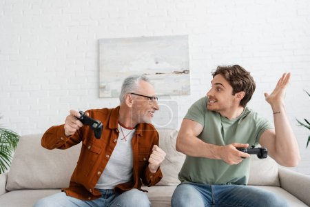 Photo for KYIV, UKRAINE - MAY 11, 2022: mature and cheerful man with joystick showing win gesture near son with displeased grimace - Royalty Free Image