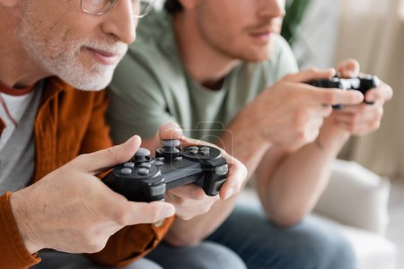 Photo for KYIV, UKRAINE - MAY 11, 2022: cropped view of mature and bearded man holding joystick while gaming with blurred son - Royalty Free Image