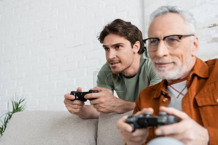 Photo for KYIV, UKRAINE - MAY 11, 2022: young and tensed man gaming with mature dad smiling on blurred foreground - Royalty Free Image