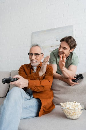Photo for KYIV, UKRAINE - MAY 11, 2022: smiling guy with joystick giving high five to mature dad playing video game on couch - Royalty Free Image