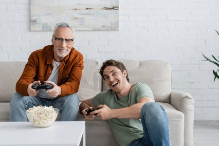 Photo for KYIV, UKRAINE - MAY 11, 2022: young excited man playing video game with mature dad sitting on couch near popcorn - Royalty Free Image