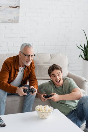 Photo for KYIV, UKRAINE - MAY 11, 2022: young astonished man gaming with mature dad smiling on sofa near popcorn - Royalty Free Image