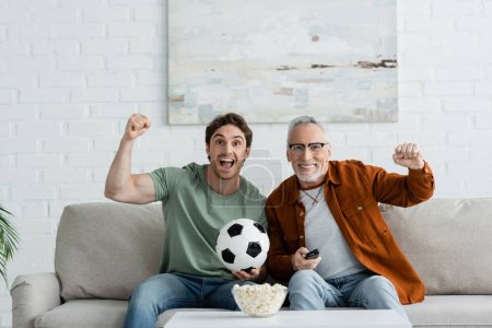 junger aufgeregter Mann mit reifem Vater zeigt Siegergeste in der Nähe von Fußball und Popcorn, während er sich die Meisterschaft im Fernsehen ansieht