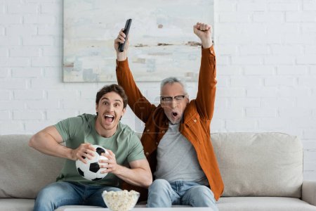 Reifer Mann zeigt Siegergeste und schreit neben aufgeregtem Sohn, der Fußballball hält, während er Fußballspiel im Fernsehen anschaut