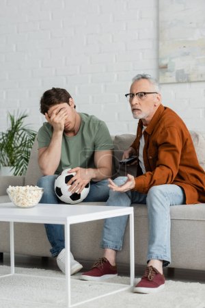 frustrated man with soccer ball obscuring face with hand near dad watching football match on tv