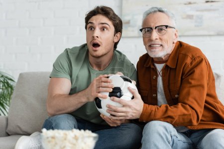 thrilled guy with tense dad holding soccer ball while watching football championship on tv