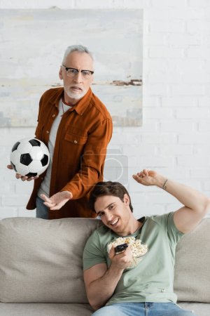 frustrated father and son with soccer ball and bowl of popcorn gesturing while watching football championship on tv
