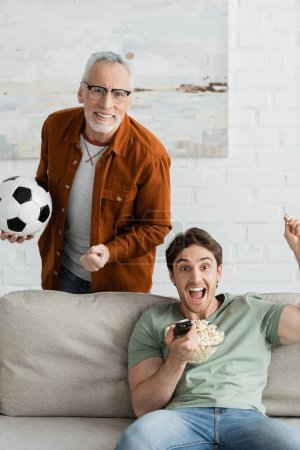 glücklicher grauhaariger Mann hält Fußballball und jubelt neben aufgeregtem Sohn mit Fernbedienung und schreit auf Couch