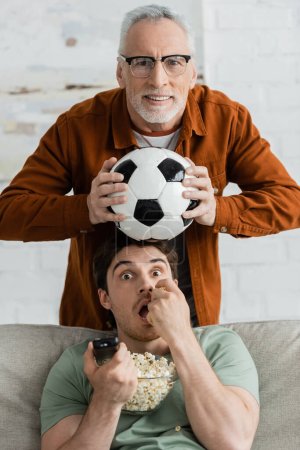 Reifer und angespannter Mann mit Fußballball beobachtet Meisterschaft in der Nähe seines kleinen Sohnes, der Popcorn isst