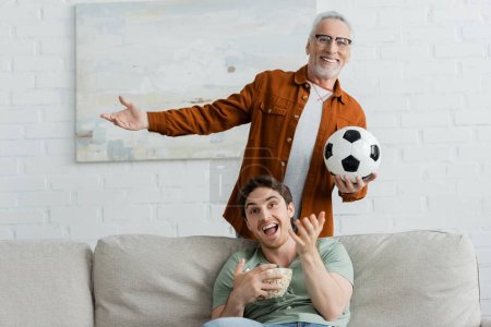 happy man with soccer ball and cheerful son with bowl of popcorn gesturing while watching football match on tv