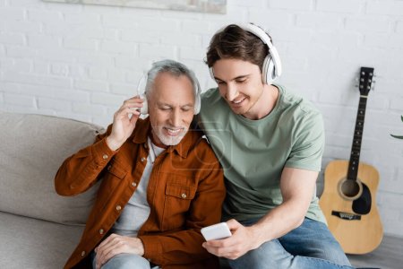 Foto de Sonriente hombre mostrando teléfono inteligente a feliz hombre de pelo gris en auriculares inalámbricos mientras escucha música en casa - Imagen libre de derechos
