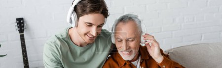 Pleased grey haired man in wireless headphones listening music near young son, banner