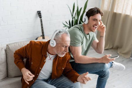 excited mature man with young son listening music in wireless headphones on couch in living room
