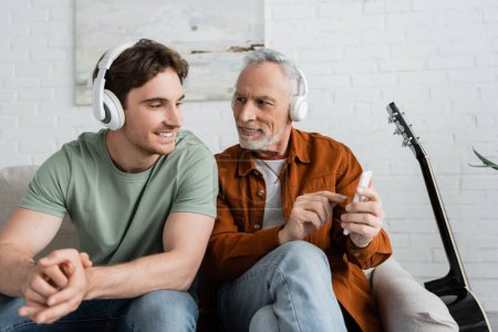 Grauhaariger Mann mit drahtlosen Kopfhörern hält Smartphone neben lächelndem Sohn 