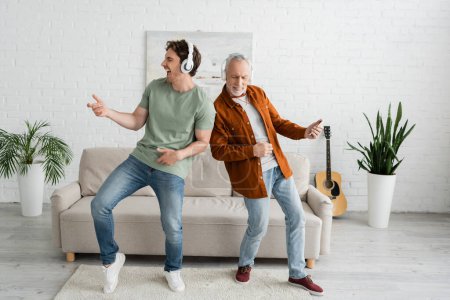 longitud completa de chico joven con padre mayor en auriculares inalámbricos escuchando música y bailando en la sala de estar
