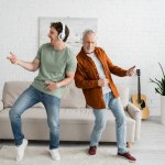 full length of young guy with senior dad in wireless headphones listening music and dancing in living room