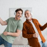 excited guy in wireless headphones singing and dancing with mature and cheerful dad at home