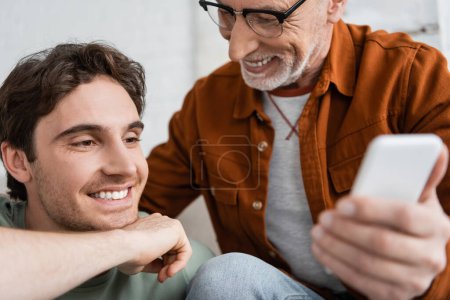glücklicher und reifer Vater mit Bart hält verschwommenes Smartphone in der Hand, während er freudigen Sohn ansieht 