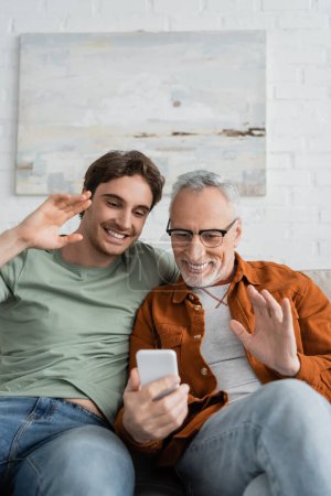 Happy mature man and cheerful son waving hands during video call on smartphone