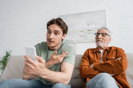 astonished young man gesturing during video call while sitting near mature father with crossed arms