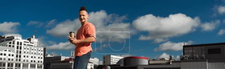 Photo for Smiling man looking at camera and holding smartphone and coffee to go on urban street, banner - Royalty Free Image