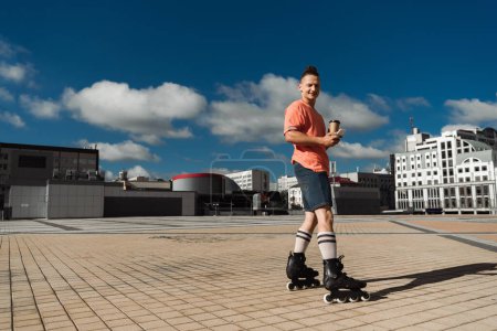 Photo for Positive roller skater holding cellphone and coffee to go while looking at camera on urban street - Royalty Free Image