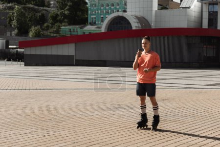 Photo for Pleased roller skater holding paper cup and mobile phone while riding on urban street - Royalty Free Image