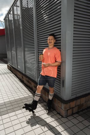 Hombre positivo en patines riendo mientras sostiene teléfono inteligente y café en la calle urbana 