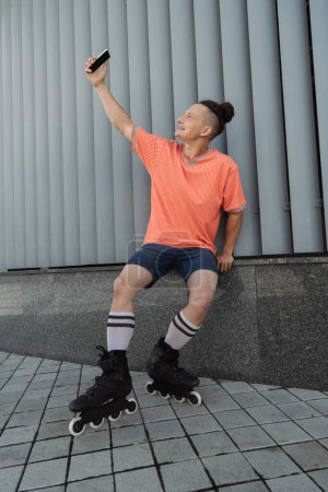 Téléchargez les photos : Vue latérale de l'homme souriant en patins à roulettes prenant selfie sur smartphone près du bâtiment à l'extérieur - en image libre de droit