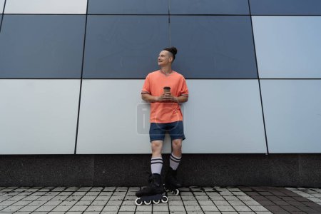 Positiver Mann auf Rollschuhen hält Pappbecher in der Nähe von Gebäuden im Freien 