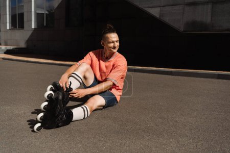 Positive man wearing rollers while sitting on asphalt on urban street 