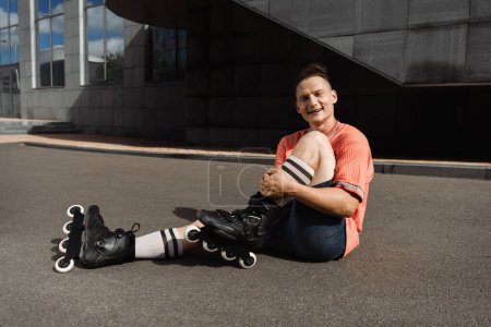 joyful and stylish roller skater sitting on asphalt and looking at camera