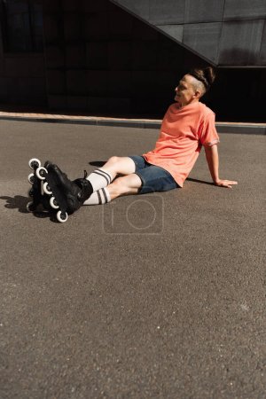 happy young roller skater with trendy hairstyle sitting on asphalt on city street