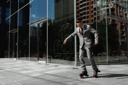 longitud completa de joven hombre de negocios en traje gris patinaje sobre ruedas cerca de edificio moderno con fachada de vidrio